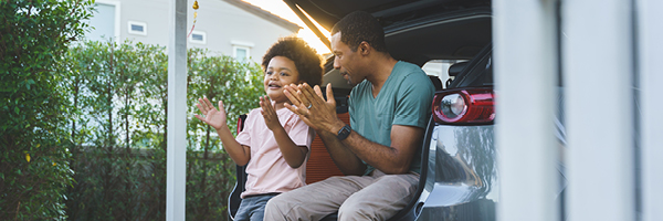 A father and son clap their hands while sitting together in the open trunk of an SUV.